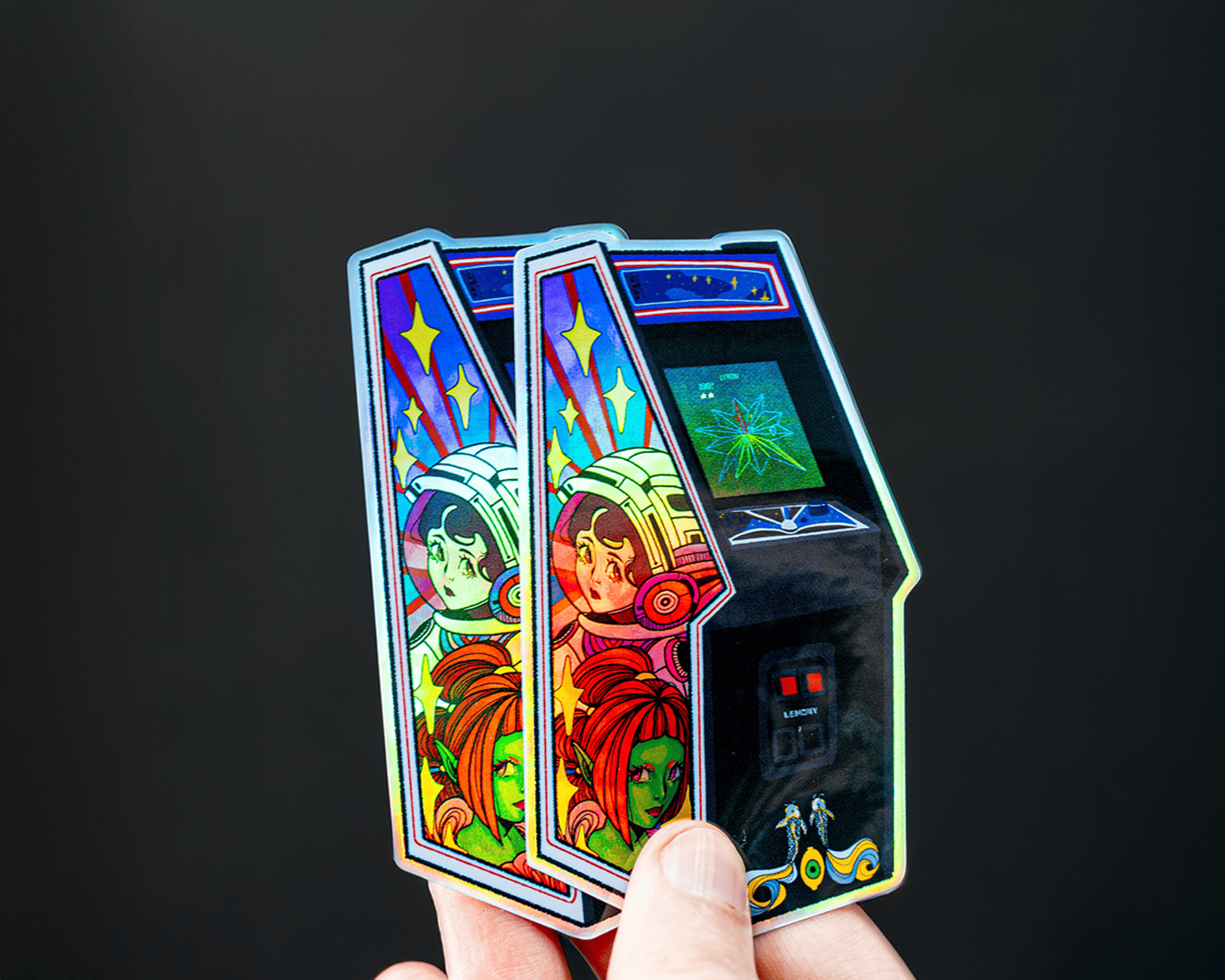 Hand holding holographic stickers of retro arcade machines with cosmic designs and astronaut illustrations against a dark background.